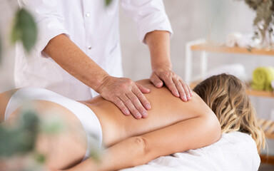 Hands of aged masseur massaging back of female patient lying on massage table