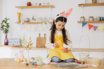 A beautiful child girl sitting on the table with decoratioons and painting colorful eggs