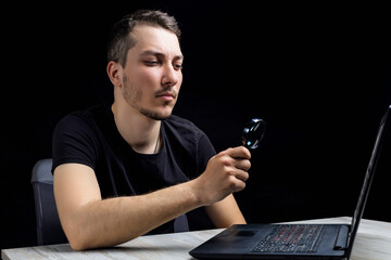 Young handsome man looks through a magnifying glass at the laptop screen on a black background....