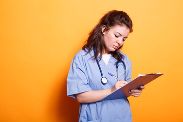 Doctor in blue scrubs writing on clipboard papers, standing against isolated orange background....