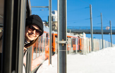 Happy woman in sunglasses sticking head out of window of mountain express train passing among snowy...