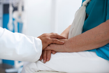 Close-up shows doctor supporting senior patient at clinic office by holding their hands. Medical professional assisting old person with chiropractic examination and diagnosis.