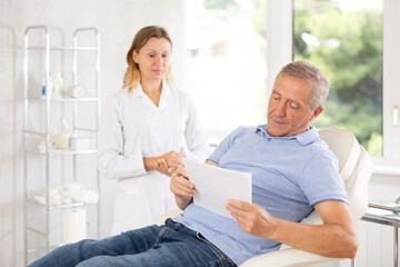 Old man patient signing documents together with female physician in light doctor's cabinet