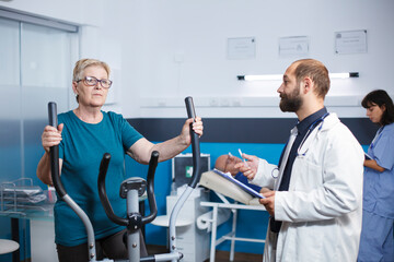 Fototapeta na wymiar Male doctor talking to senior patient doing physical activity to recover from injury with electrical bike. Senior woman using stationary bicycle for wellness and recovery at physiotherapy clinic.