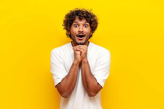 shocked excited curly indian guy in white t-shirt with open mouth looking at camera on yellow isolated background, surprised guy in amazement