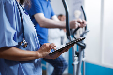 Close-up of physician reviewing healthcare report on tablet while wearing blue scrubs and stethoscope at a clinic. Caucasian nurse practitioner browsing the internet on digital device.