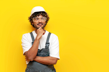 young pensive indian male builder in hard hat and overalls plans and thinks on yellow isolated...