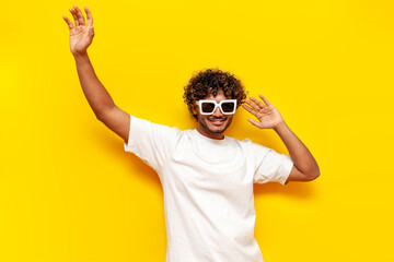 young cheerful indian man in white t-shirt and sunglasses dancing with raised hands on yellow...