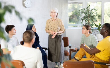 Friendly and calm senior female psychotherapist or facilitator leading group therapy session with women and men of different ages and nationalities sitting 