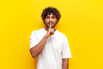 young indian man in a white t-shirt holds his finger near his lips and shows a hush gesture on a yellow isolated background, curly-haired guy asks to be silent and hides a secret