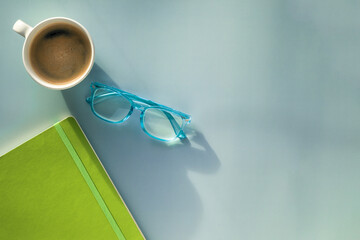 A morning coffee mug, a pair of blue reading glasses and a green notebook on a solid blue background