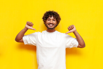 young confident indian man in white t-shirt pointing to himself and smiling on yellow isolated...