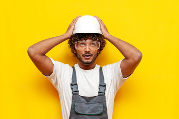 A young Indian builder in safety glasses and overalls puts a hard hat on his head on a yellow...