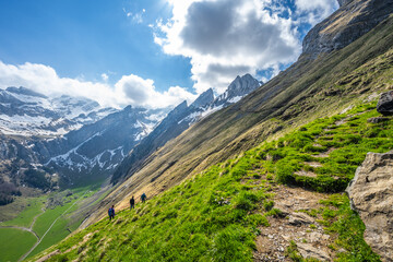 Tourists walk along a hiking trail through a green meadow with an alpine valley and a steep, rocky mountain formation in the background. Seealpsee, Appenzell, Switzerland, Europe.