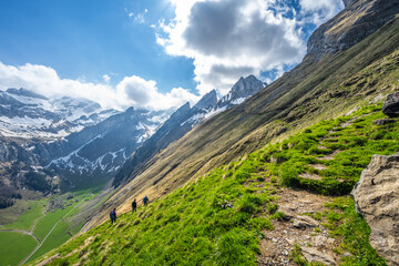 Tourists walk along a hiking trail through a green meadow with an alpine valley and a steep, rocky...