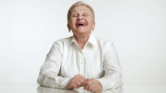 Funny intelligent woman of 60-70 years sitting in white studio at white table raising head, laughing with mouth open, humorous elderly teacher pranking. High quality 4k footage