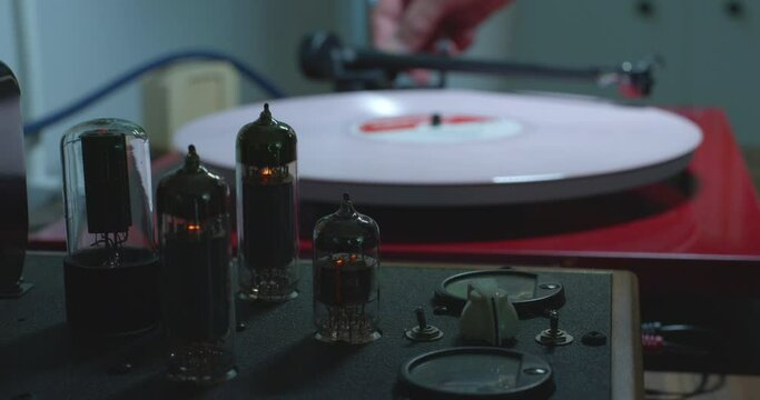 Close up of tube amplifier with turntable in the background with man's hand placing a stylus on pink colored vinyl record.