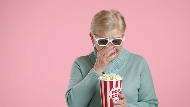 Senior lady with blonde short hair in white-rimmed 3d glasses standing over pink background sobbing, wiping tears with hand, sympathizing with grief. High quality 4k footage