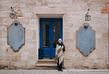 A young pretty girl in an autumn coat, scarf and hat stands near the blue door of a cafe on the street of Lviv, Ukraine.