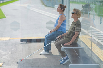 An elderly blind woman and a mature pregnant woman are sitting at a bus stop and waiting for the...