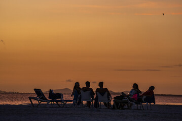 Grupo de personas sentadas conversando mientras observan el atardecer en la playa de una isla en...