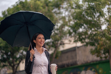 A beautiful Latin woman walks down the street on a rainy day holding an umbrella and a coffee in...
