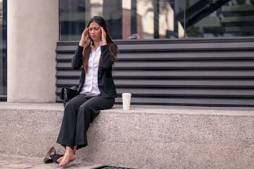 Business woman sitting on the street with headache. High shoes and a formal suit massages her head.