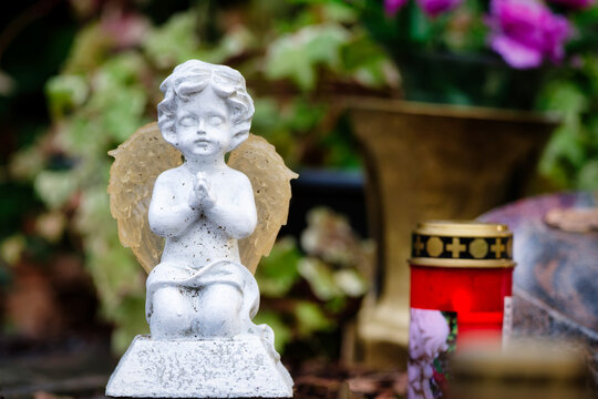 a small white angel figure kneels praying on a grave in front of a grave candle and gravestone as well as flowers in the blurred background