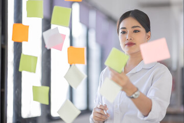 Business Asian woman meeting at office and use post it notes in glass wall to share idea. Brainstorming business office concept. Sticky note on glass wall.
