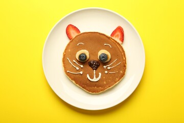 Creative serving for kids. Plate with cute cat made of pancakes, berries, cream, banana and chocolate paste on yellow background, top view