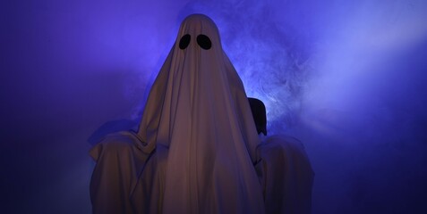 Creepy ghost. Woman covered with sheet sitting in armchair in blue light