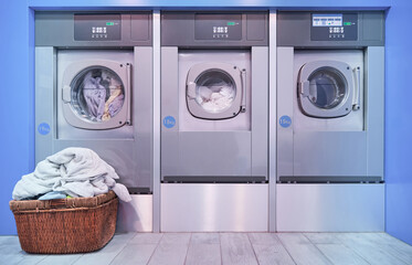 Basket full of dirty clothes in front of three industrial washing machines operating inside a self...