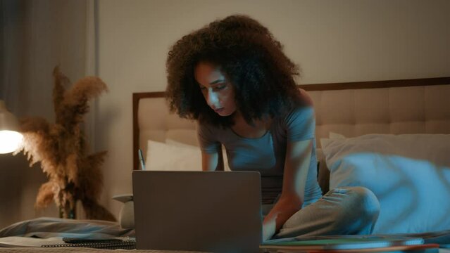 African American 20s girl high school student learning at home writing notes homework on bed late night online studying with laptop distant education young woman listen webinar on computer e-learning