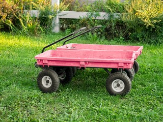 Two well-worn flat red plastic gardening or toy wagons side by side in a yard with green grass, in front of a white wood fence and wild growing bushes. A bit of leaves peek out from one of the wagons.