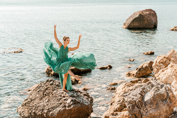 Woman green dress sea. Female dancer in a long mint dress posing on a beach with rocks on sunny day. Girl on the nature on blue sky background.