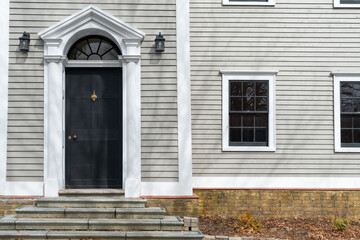 The exterior of a vintage building with beige-colored narrow clapboard Cape Cod siding. There's a black wooden door with a thick white decorative trim. A half-circle transom window hangs over the door - obrazy, fototapety, plakaty
