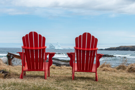 Two vibrant red empty Adirondack chairs face the Atlantic Ocean and cloudy blue sky on a grassy coastline. There's a large white glacier iceberg floating in the distance between the two chairs.   