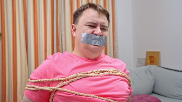 distressed Man in pink shirt grimacing tied with ropes and mouth sealed with duct tape, hinting at an abduction scenario in a domestic setting