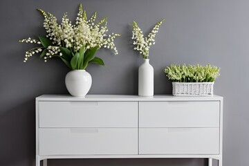 White modern dresser minimalistic furniture in empty room on grey wall background, small cupboard with decor, vases and lily of the valley flowers bouquet, cozy apartment house interior.