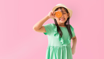 Happy Asian little girl posing with wear a hat with sunglasses holding orange slices on face, Holiday summer fashion green dress, isolated on pastel pink color background - obrazy, fototapety, plakaty