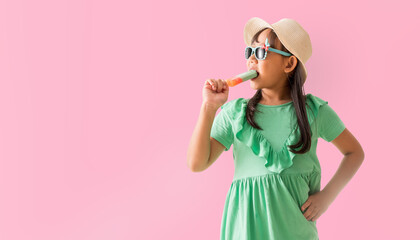 Happy Asian little girl posing with wear a hat with sunglasses holding ice cream, Holiday summer fashion green dress, isolated on pastel pink color background