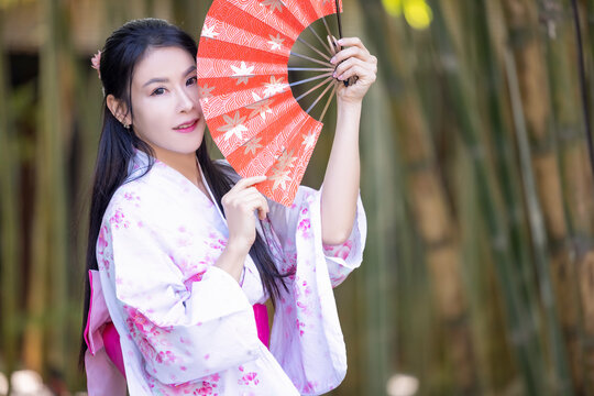 Asian woman wearing japanese traditional kimono standing on street. Beautiful Woman smiling and holding hand fan to cover face. Woman in Kimono dress enjoying and walking on bamboo forest.