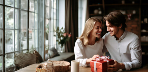young cheerful couple is sitting in a cafe at a table near a large window. Red surprise gift box for a girl. Free space for text. Valentine's Day, 14.02, New Year