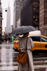 A girl in a gray coat with a black umbrella stands on a city street waiting for a taxi