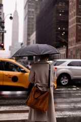 A girl in a gray coat with a black umbrella stands on a city street in front of a pedestrian crossing and wants to cross the road. The rainy city is full of cars.