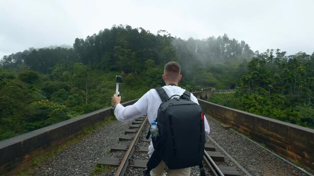 Rear view of a man tourist taking pictures of mountain forested landscape on smartphone. Action. Hiker walking on railways.