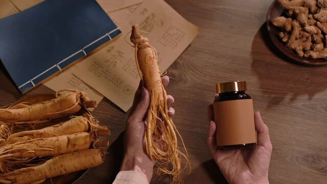 Top view of one hand holding a ginseng root, the other holding an unlabeled medicine bottle. A plate of ginseng, ancient books and a plate of medicinal herbs are displayed on a wooden table.