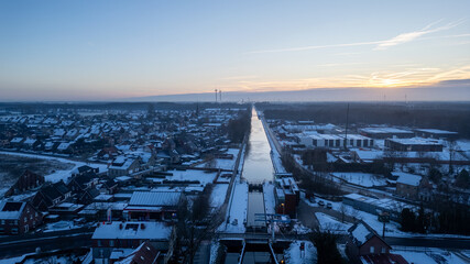This aerial image presents a serene winter morning over a suburban area, with a canal cutting through the heart of the neighborhood. The sunrise brings a warm glow to the cool blue tones of the snow
