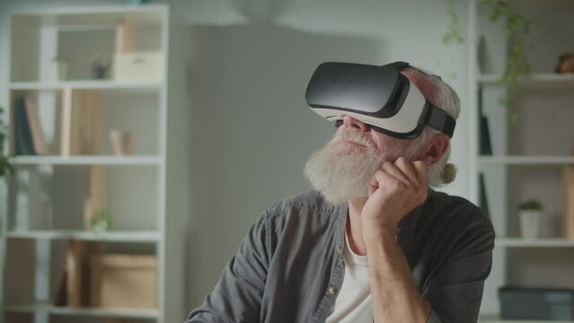 A Modern Old Man in VR Glasses Reads a Virtual Book. An Elderly Man in VR Glasses Sitting at a Table at Home and Examines Everything Around.Virtual Reality Education for the Elderly.