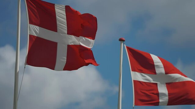 Two Danish flags flutter in the wind in the slow-motion video. Blue sky with white clouds in the background.